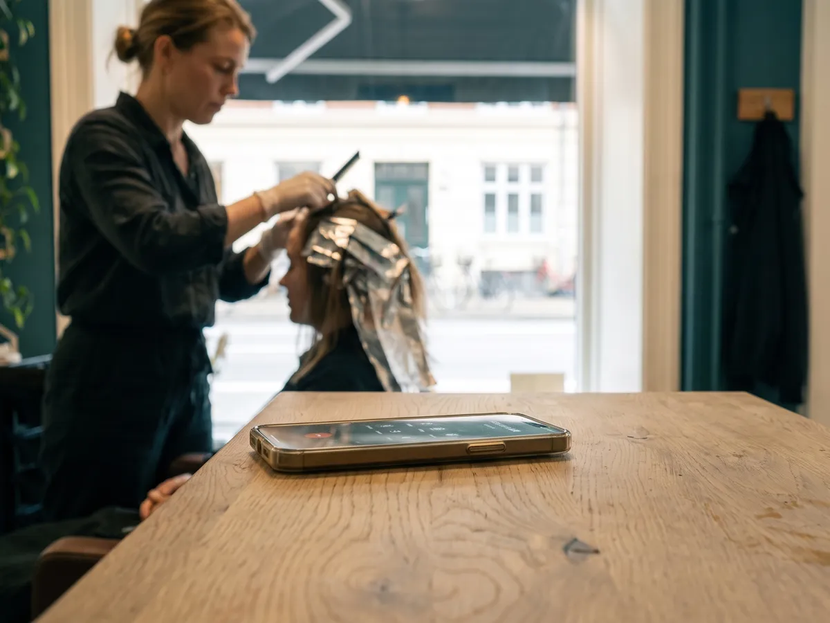 En frisør lægger folie i en kundes hår. Telefonen ringer på disken.